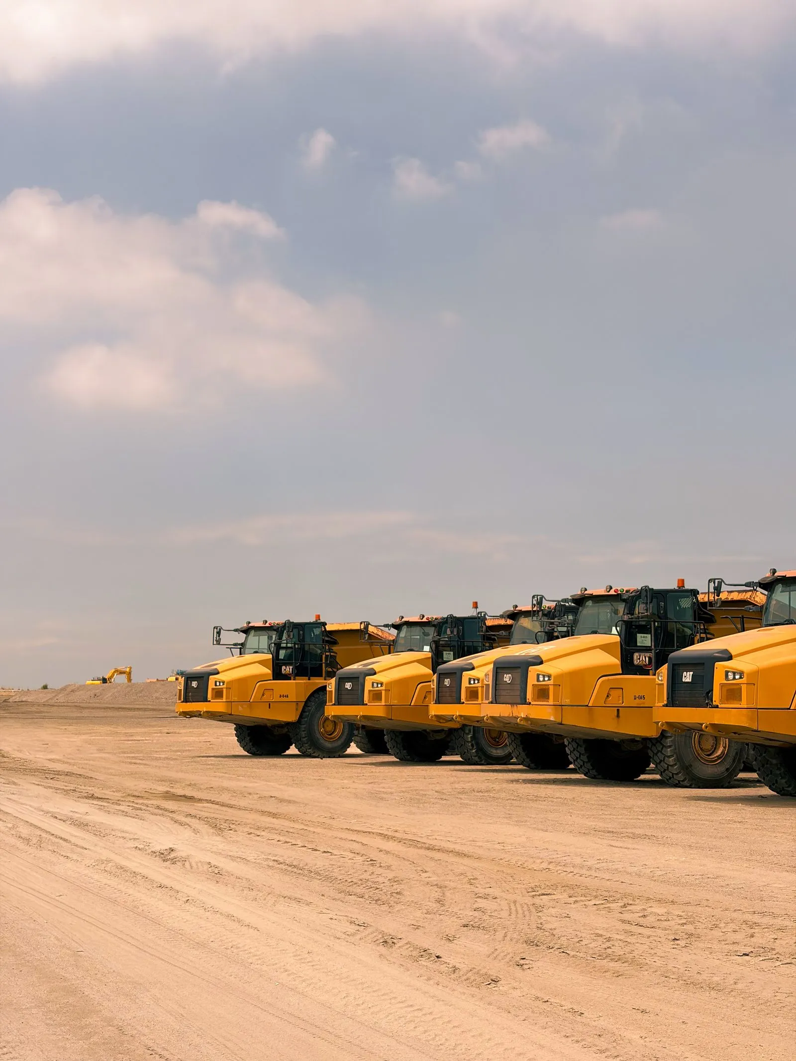 A row of yellow articulated dump trucks parked on a flat, sandy job site